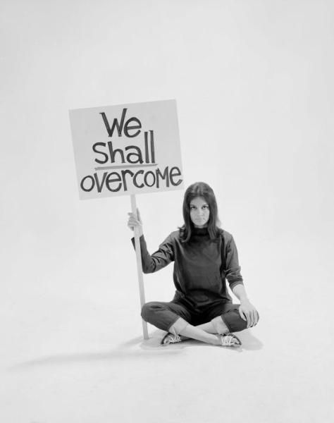 Writer Gloria Steinem sitting cross-legged on floor w. sign "We Shall Overcome" re pop culture